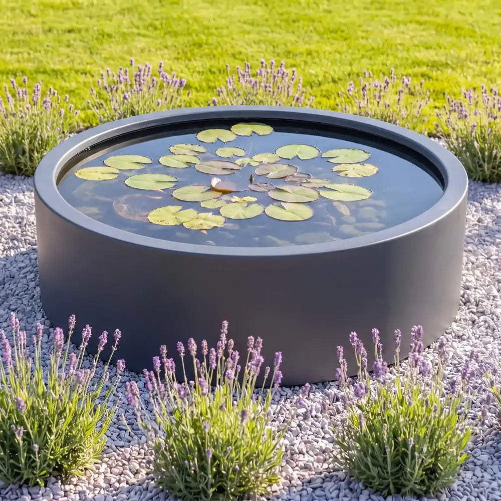 Rundes Alu-Wasserbecken mit Wasserpflanzen auf einer Kiesfläche in einem minimalem Gartendesign.