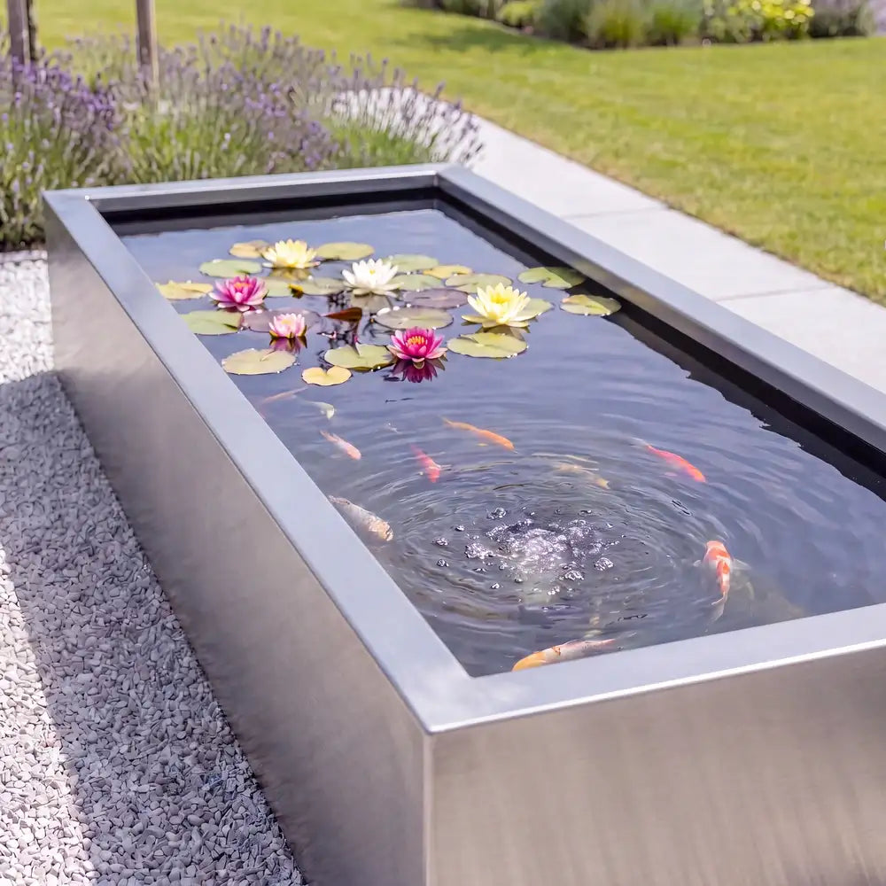 Rechteckiges Wasserbecken aus Edelstahl mit Seerosen und Fisch auf einer Kiesfläche in einem modernen Garten.