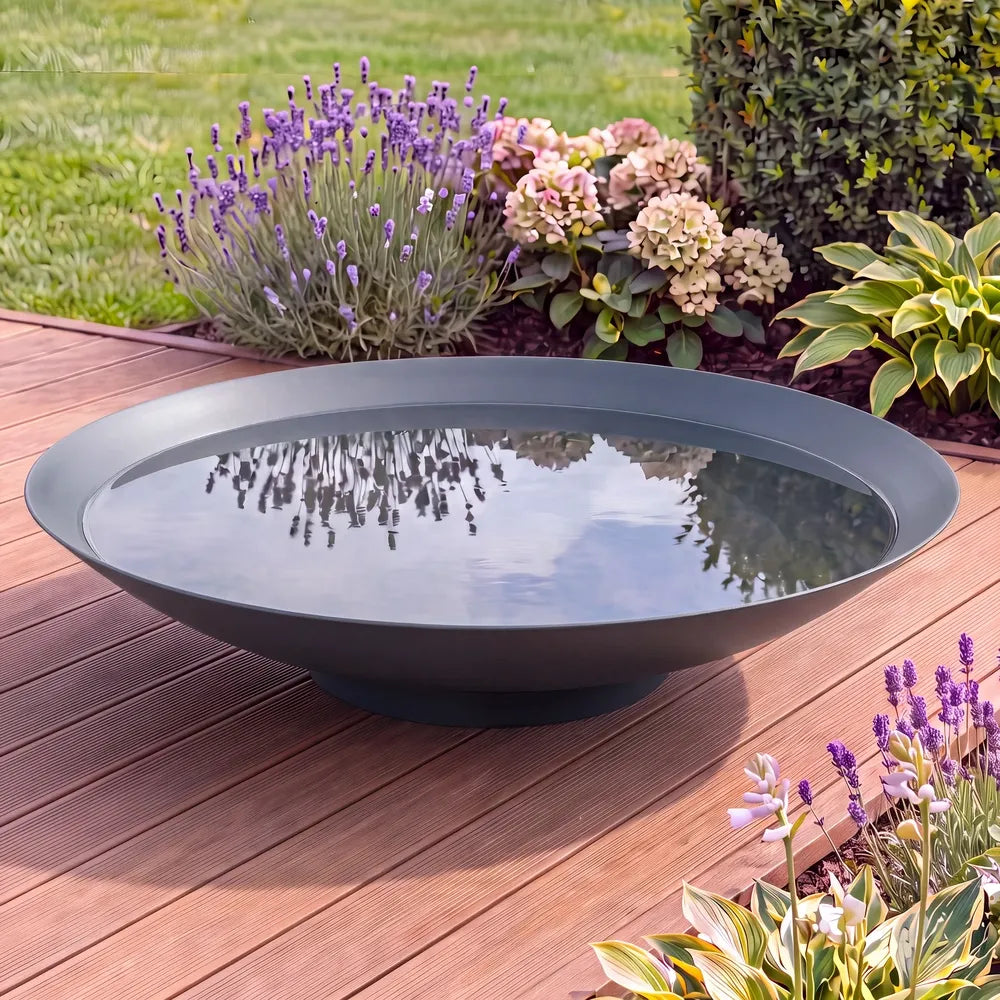 Runde Metall-Wasserschale auf einer modernen Terrasse in einem Garten und Blumen im Hintergrund.