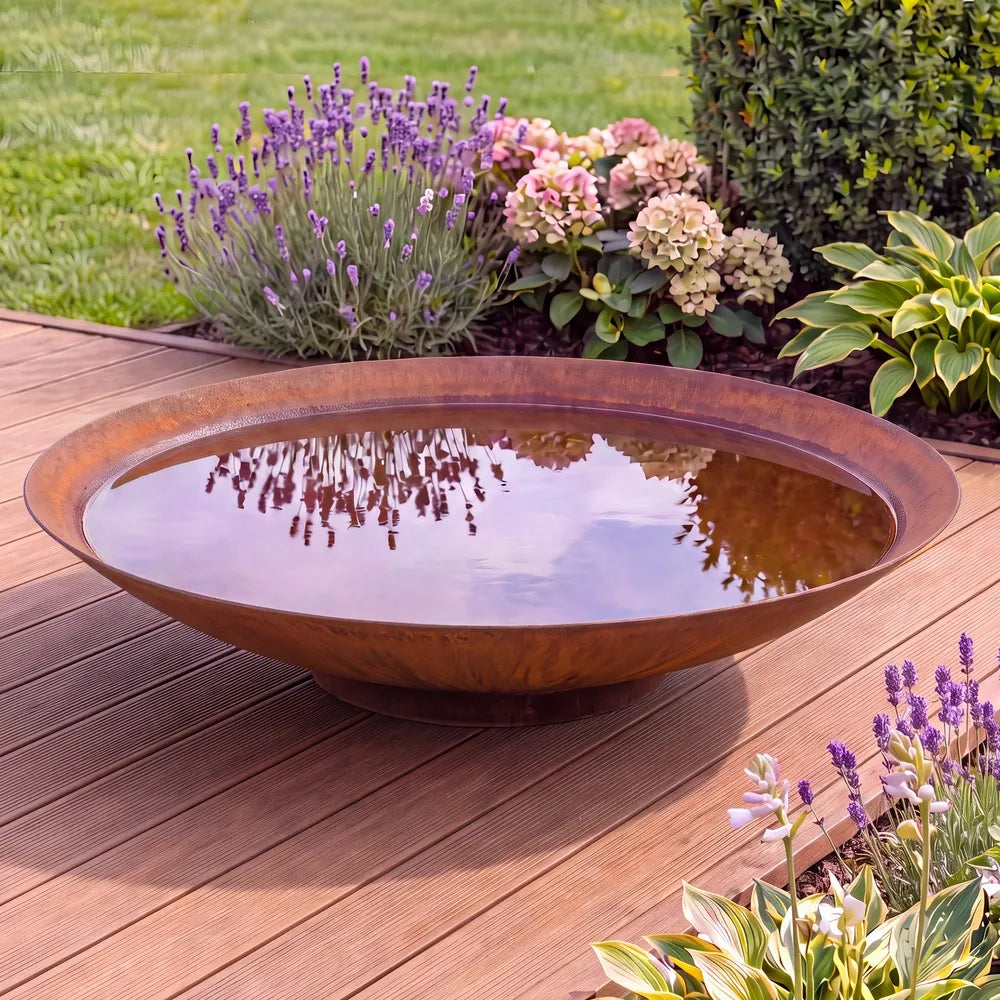 Runde Cortenstahl-Wasserschale auf einer modernen Terrasse in einem Garten und Blumen im Hintergrund.
