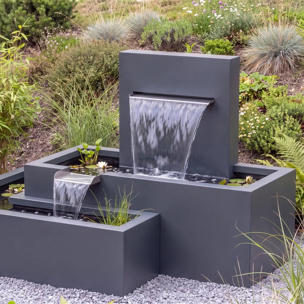 Aluminium-Kaskadenbrunnen mit versetzten Metall-Wasserbecken auf einer Kiesfläche vor einem begrünten Abhang.