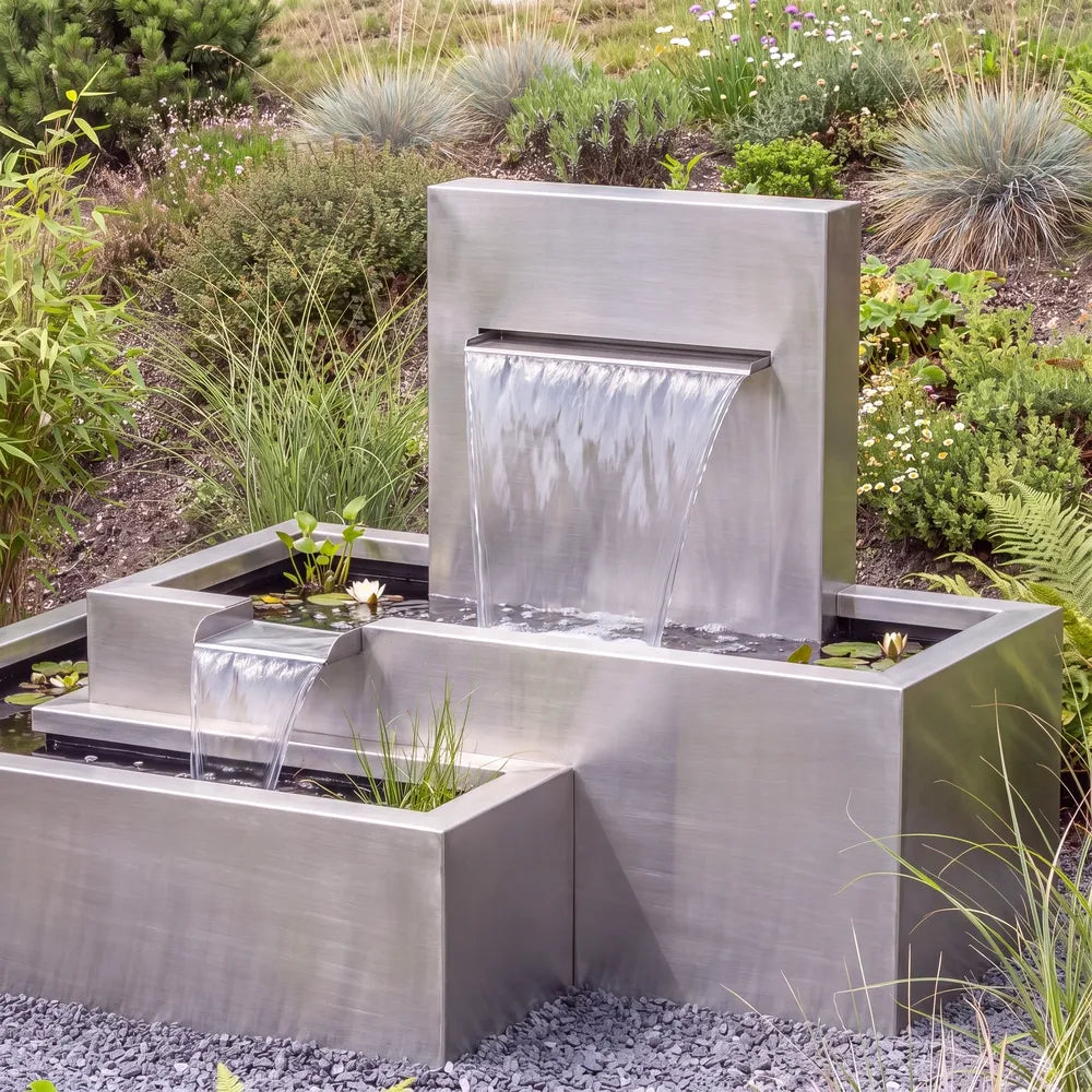 Edelstahl-Kaskadenbrunnen mit versetzten Metall-Wasserbecken auf einer Kiesfläche vor einem begrünten Abhang.