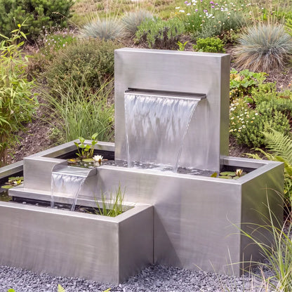 Edelstahl-Kaskadenbrunnen mit versetzten Metall-Wasserbecken auf einer Kiesfläche vor einem begrünten Abhang.