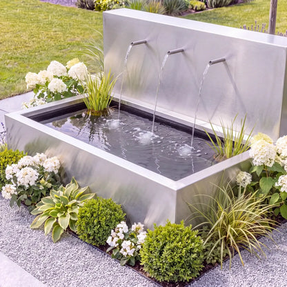 Moderne Teichwand aus Edelstahl mit Edelstahl-Einlaufrohren an einem Metall-Wasserbecken auf einer Kiesfläche mit Pflanzen und Blumen.