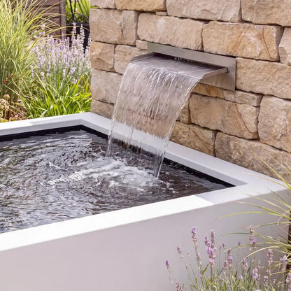 Quadratisches Wasserbecken aus Aluminium vor einer Natursteinmauer mit einer Edelstahl-Wasserschütte in einem Pflanzbeet.