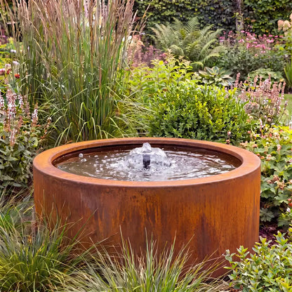 Rundes Corten-Teichbecken in einem Garten als Gartenbrunnen mit flacher Wasserfontäne.