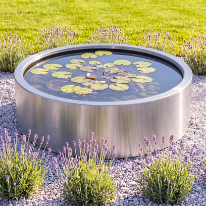 Rundes Edelstahl-Wasserbecken mit Wasserpflanzen auf einer Kiesfläche in einem minimalem Gartendesign.