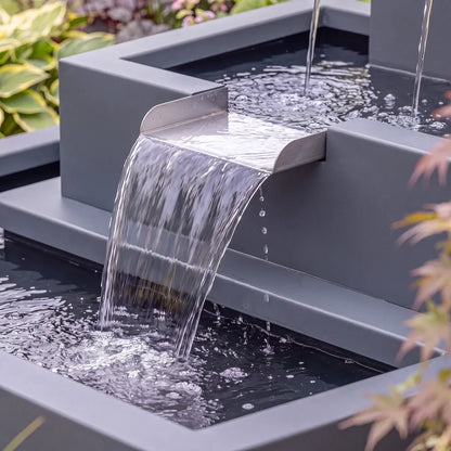 Wasserkaskade aus Aluminium mit drei Wasserausläufen und Wasserschütte in einem grünen Blumenbeet.