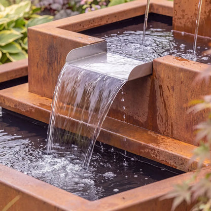 Wasserkaskade aus Cortenstahl mit drei Wasserausläufen und Wasserschütte in einem grünen Blumenbeet.