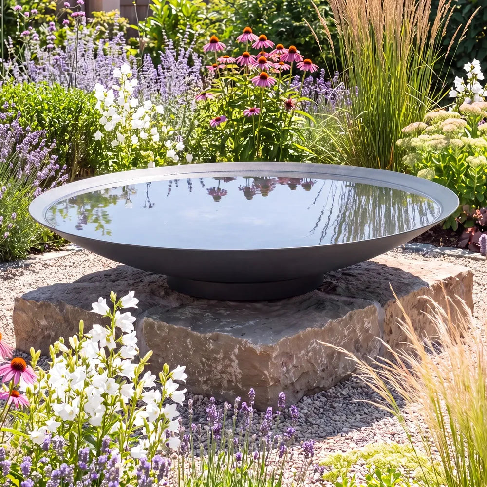 Große runde Wasserschale aus Metall mit Wasser auf einem Podest im einem bunten Blumenbeet.