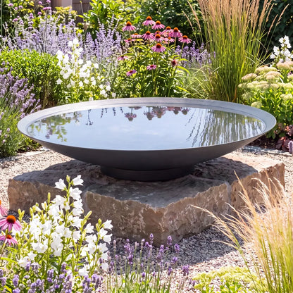 Große runde Wasserschale aus Metall mit Wasser auf einem Podest im einem bunten Blumenbeet.