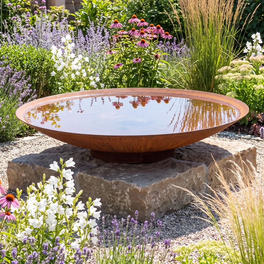Große runde Wasserschale aus Cortenstahl mit Wasser auf einem Podest im einem bunten Blumenbeet.