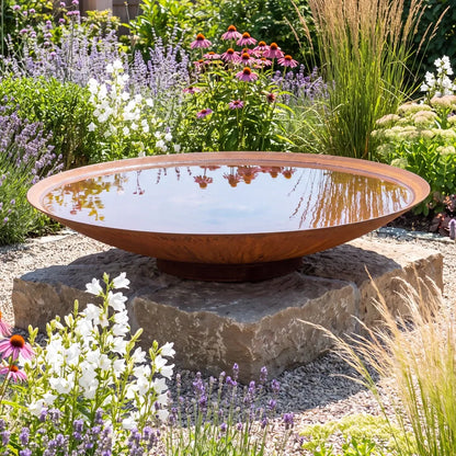 Große runde Wasserschale aus Cortenstahl mit Wasser auf einem Podest im einem bunten Blumenbeet.