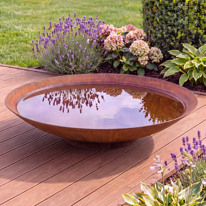 Runde Cortenstahl-Wasserschale auf einer modernen Terrasse in einem Garten und Blumen im Hintergrund.